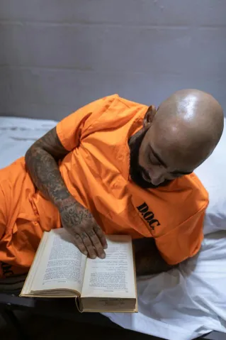 A bald, tattooed prisoner reading a book while lying on a bed in his cell.